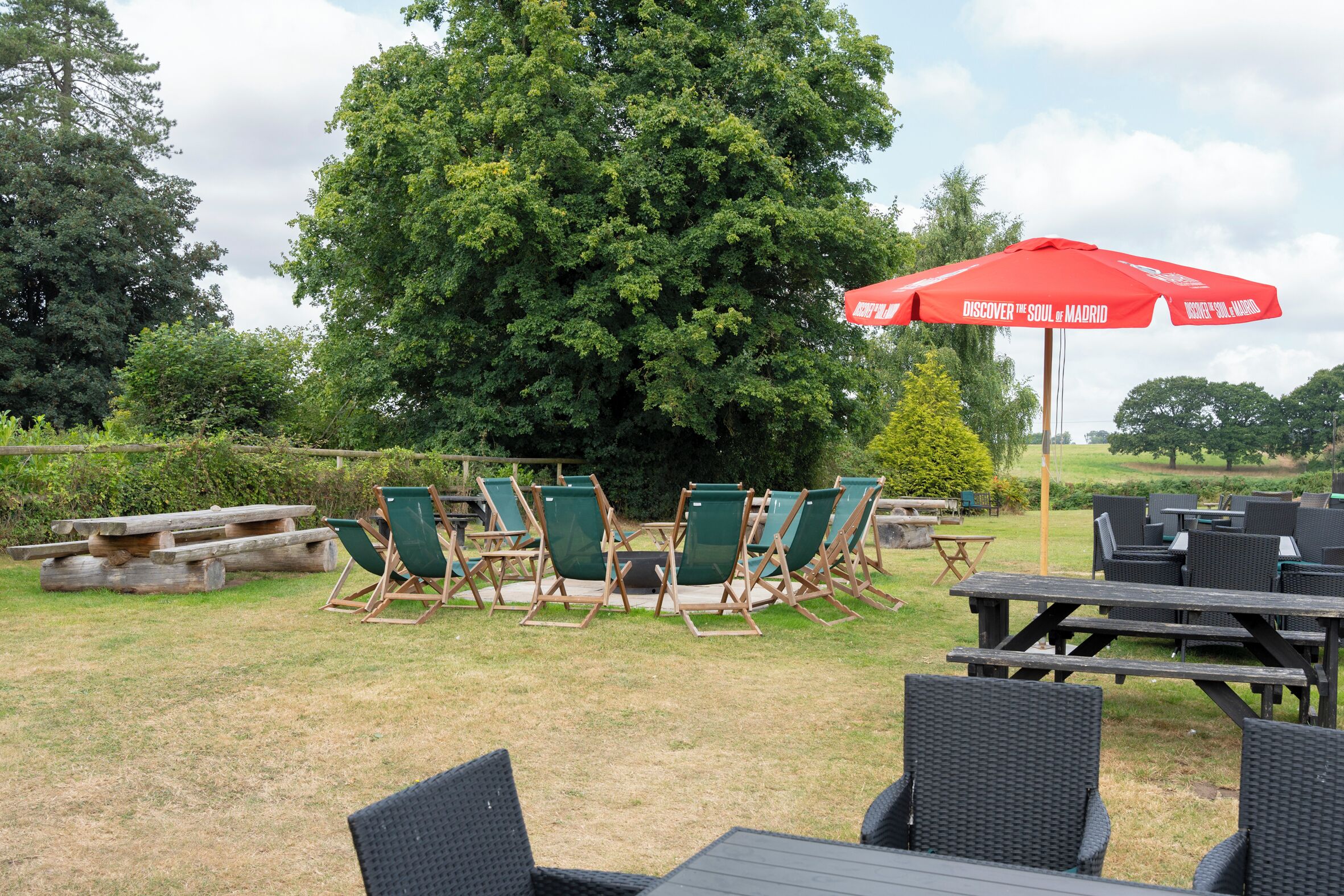 An outdoor seating area with green lawn chairs and tables, a red umbrella, and a large tree in the background.