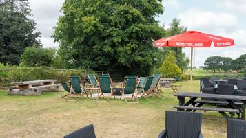 An outdoor seating area with green lawn chairs and tables, a red umbrella, and a large tree in the background.