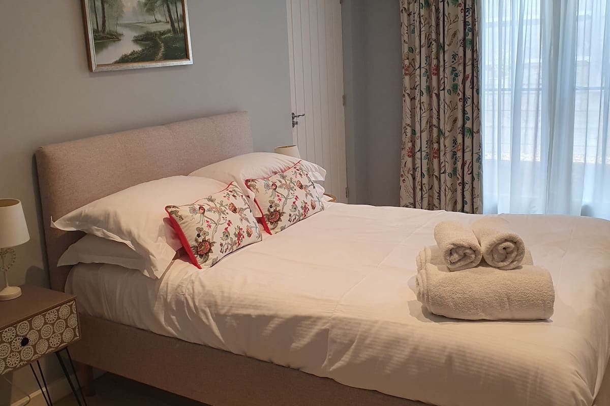 A neatly made bed with white linens and floral pillows, a painting of a forest scene on the wall, and a window with floral curtains.