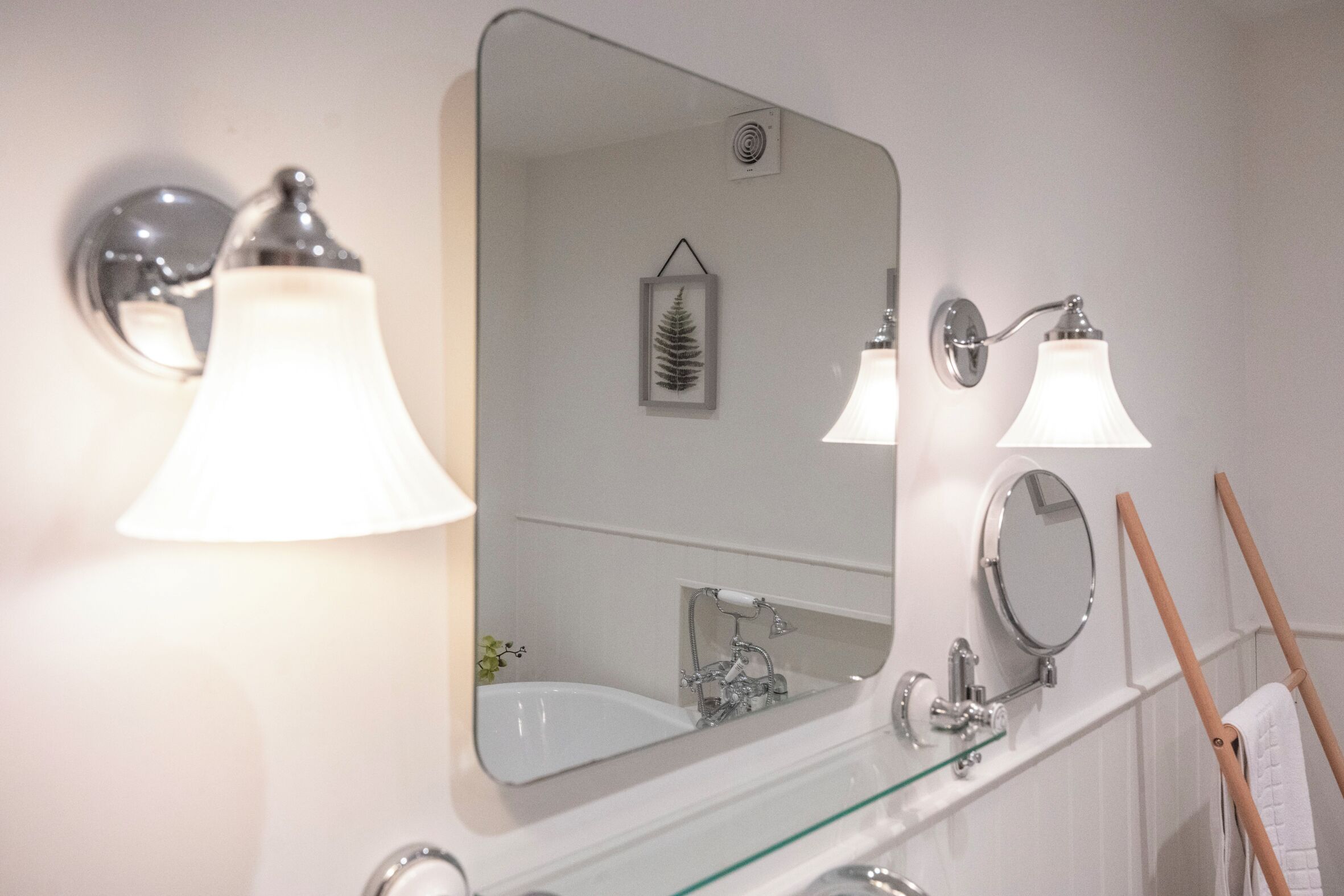 A bathroom with a large mirror, two wall-mounted lights, a glass shelf, and a towel rack.