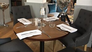 Uma sala de jantar com uma mesa redonda posta para seis pessoas, rodeada por cadeiras estofadas com detalhes metálicos.
