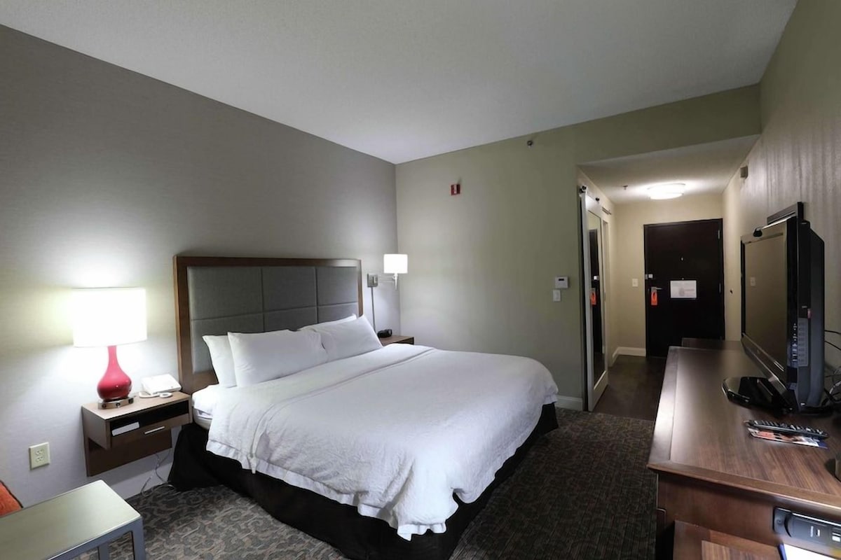 Habitación de hotel con cama, mesita de noche, lámpara, televisión y escritorio con silla.