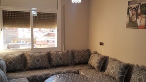 A living room with a sofa set, a chandelier, and a window with blinds. - Appartement De Luxe 4 Chambres (Oujda)