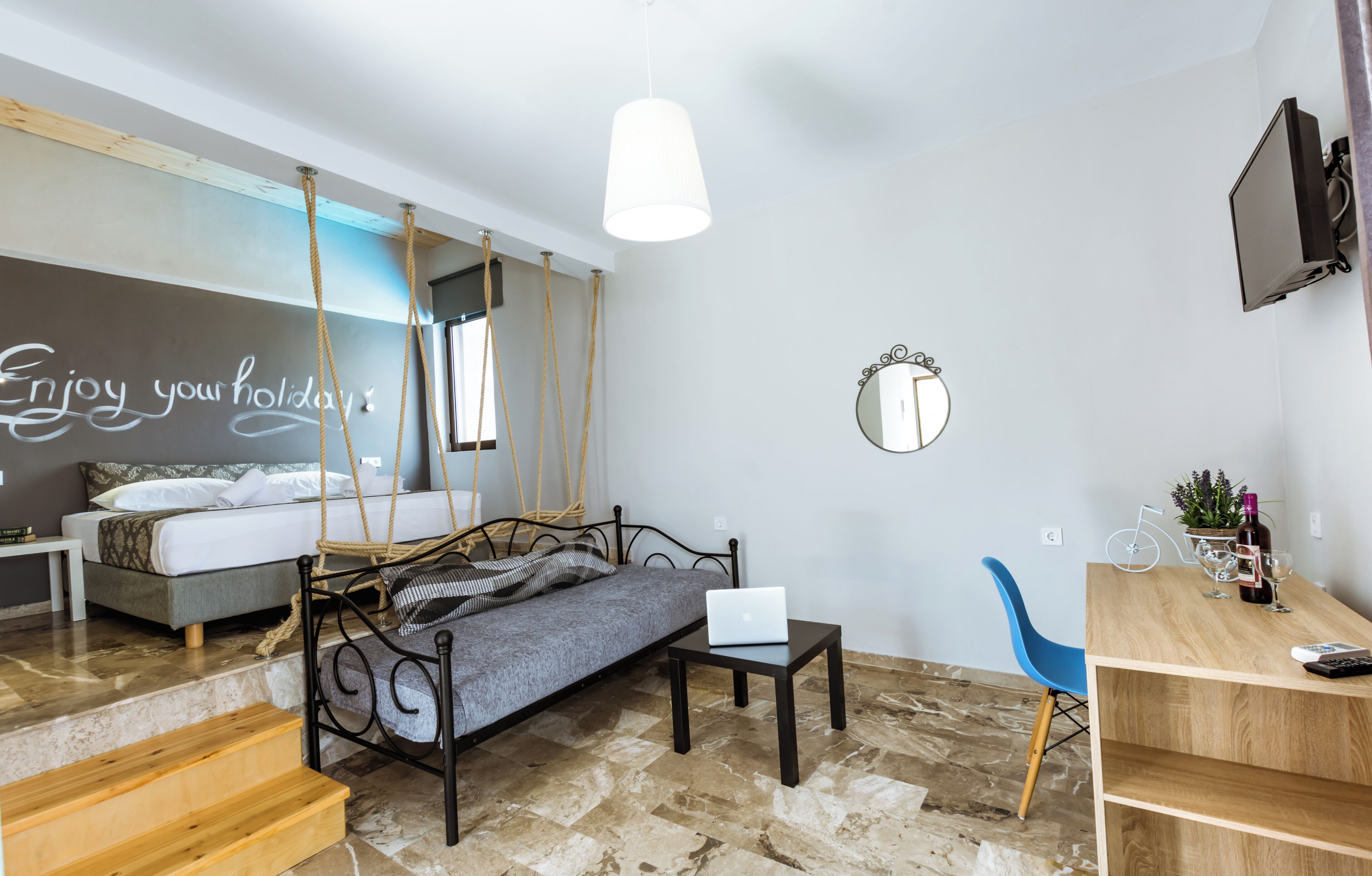 Un dormitorio moderno con una cama, un ordenador portátil sobre una mesita, un sillón colgante y un escritorio con un televisor.