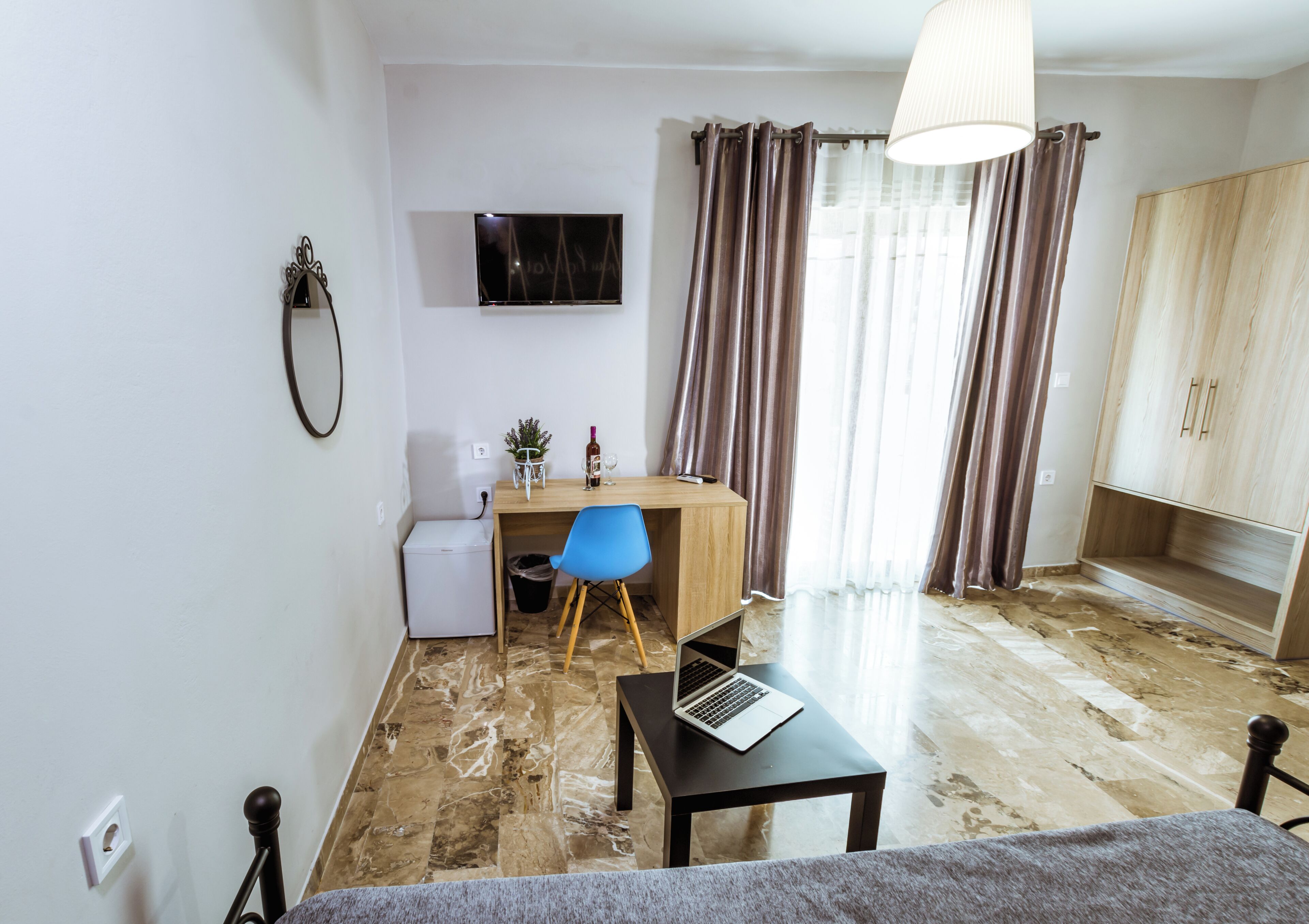 A small, modern room with a desk, chair, and a laptop. There is a television mounted on the wall, a small refrigerator, and a cabinet.