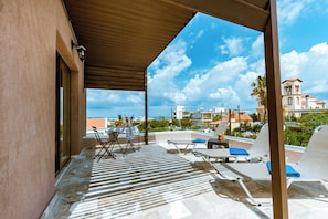 A covered outdoor area with lounge chairs, a table with chairs, and a view of buildings and palm trees. - Gregory Comfort Hotel (Chania)