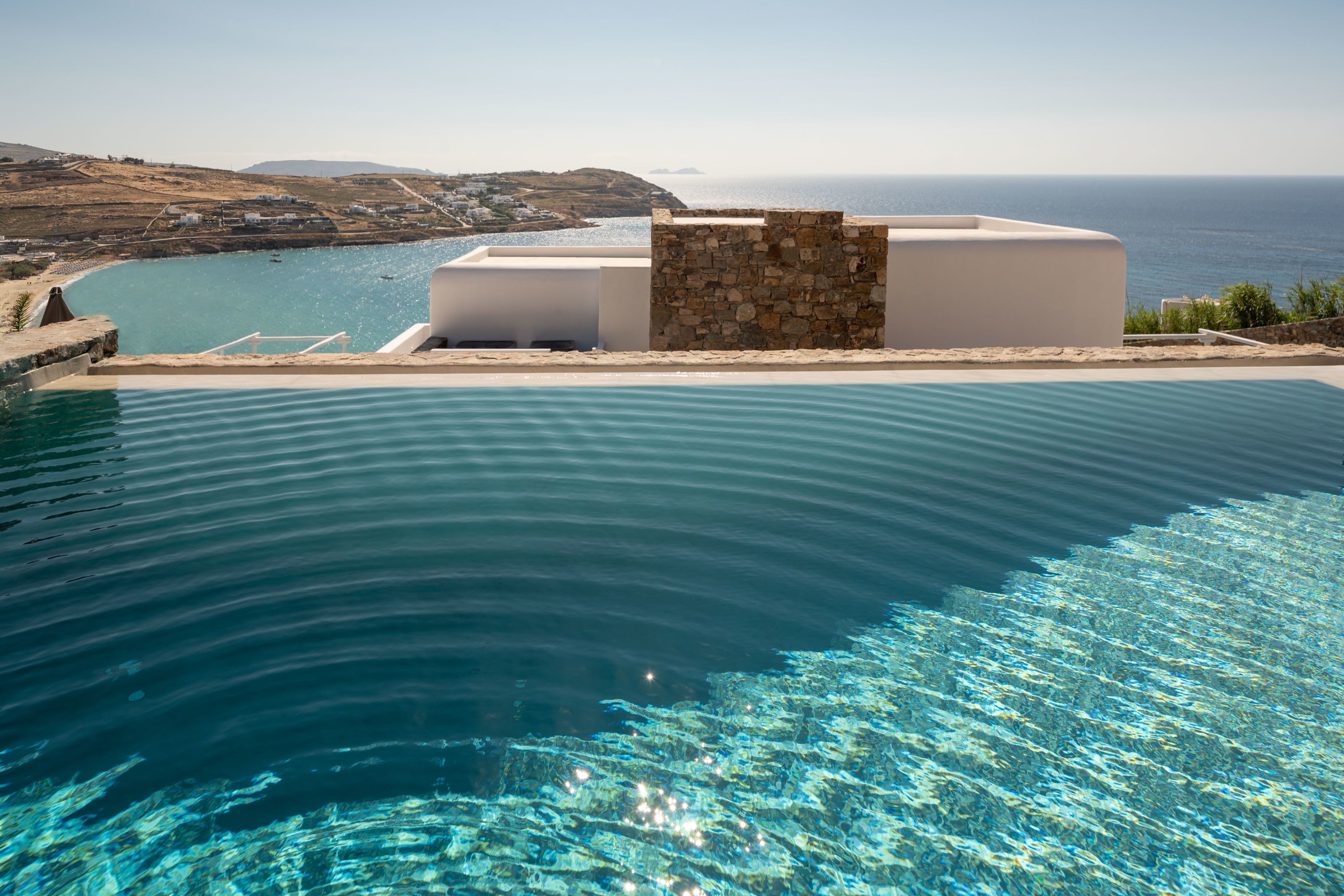 Ein Infinity-Pool mit klarem, blauem Wasser, einer Steinsäule und Blick auf das Meer und das entfernte Land.