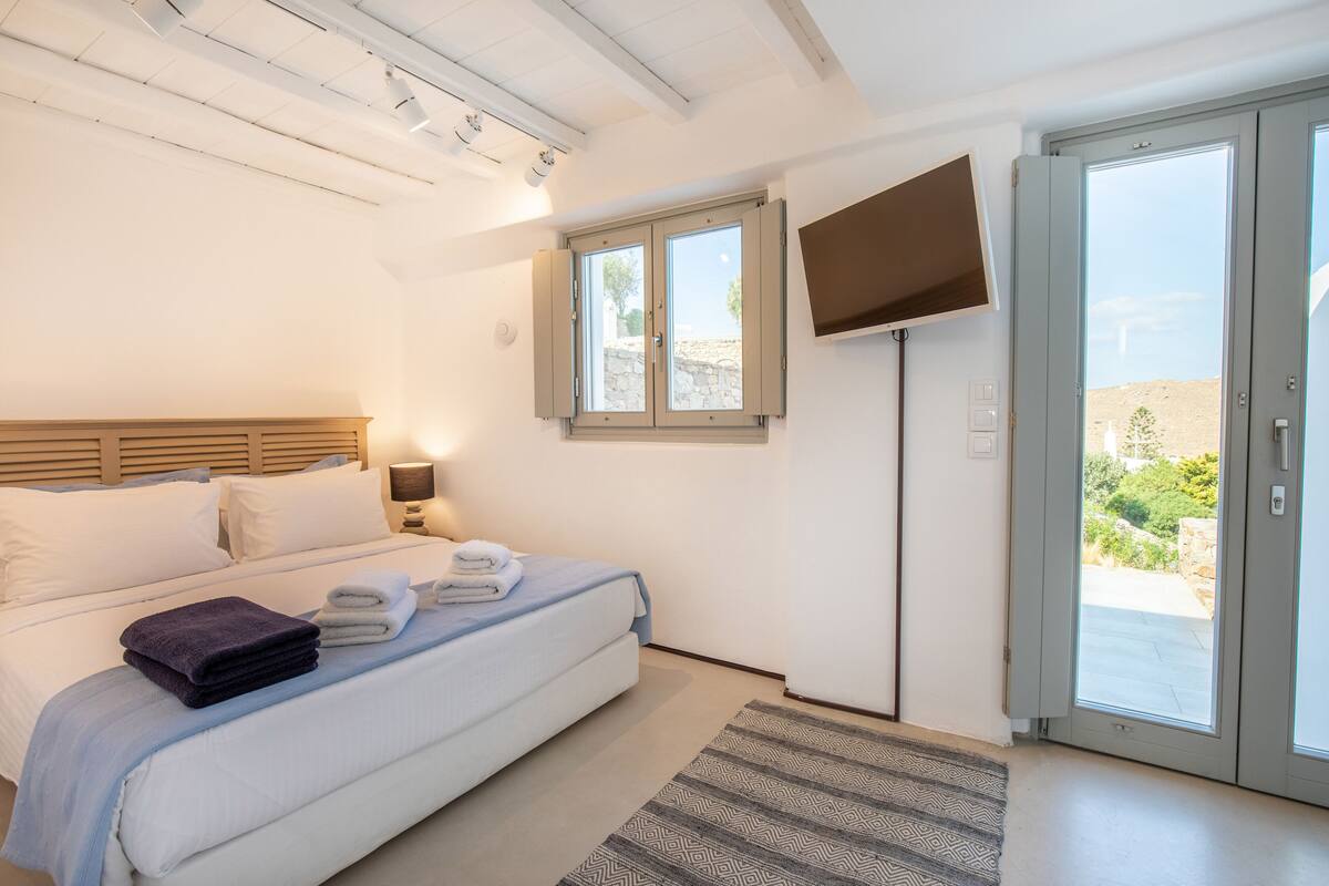 A bedroom with a bed, a TV mounted on the wall, a window, and a door leading to an outdoor area.