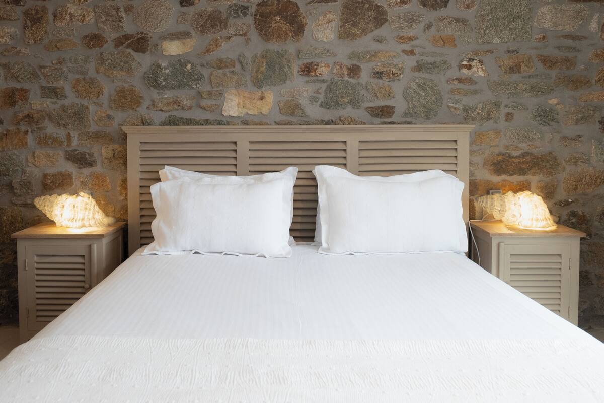A bed with white linens and a wooden headboard, flanked by two bedside tables with lamps, set against a stone wall.