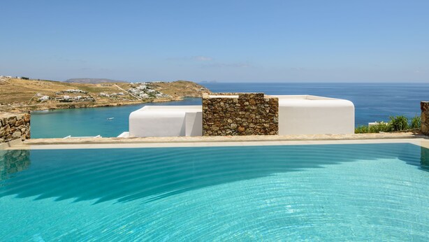 Piscina azul cristalina con un muro de piedra y un edificio blanco al fondo, con vistas a un paisaje costero.