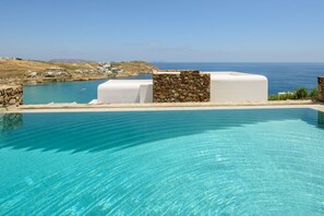 Piscina azul cristalina con un muro de piedra y un edificio blanco al fondo, con vistas a un paisaje costero.