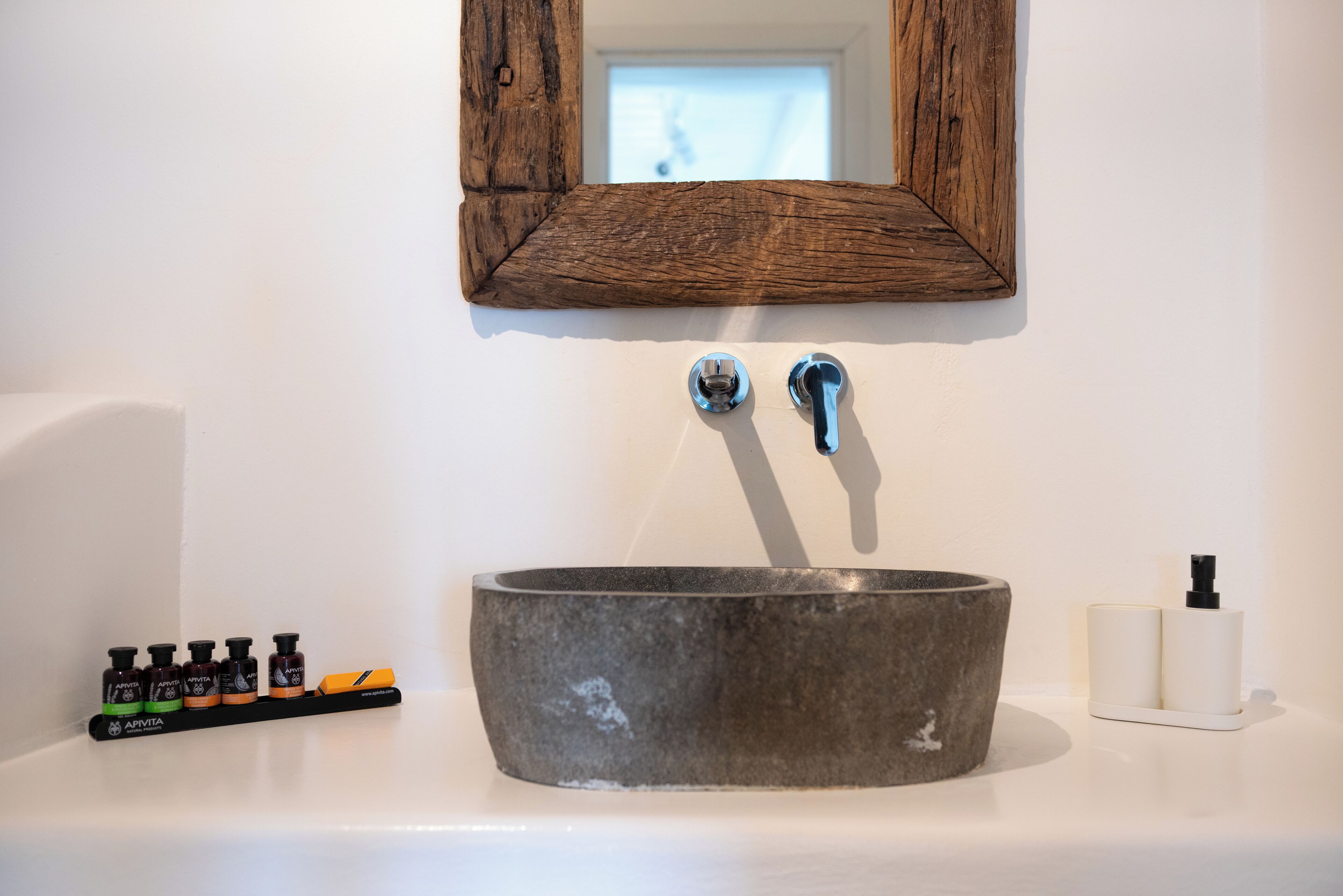 Een badkamer met een stenen wastafel, twee kraanwerkstukken, een spiegel met houten frame en een rij toiletartikelen op het aanrecht.