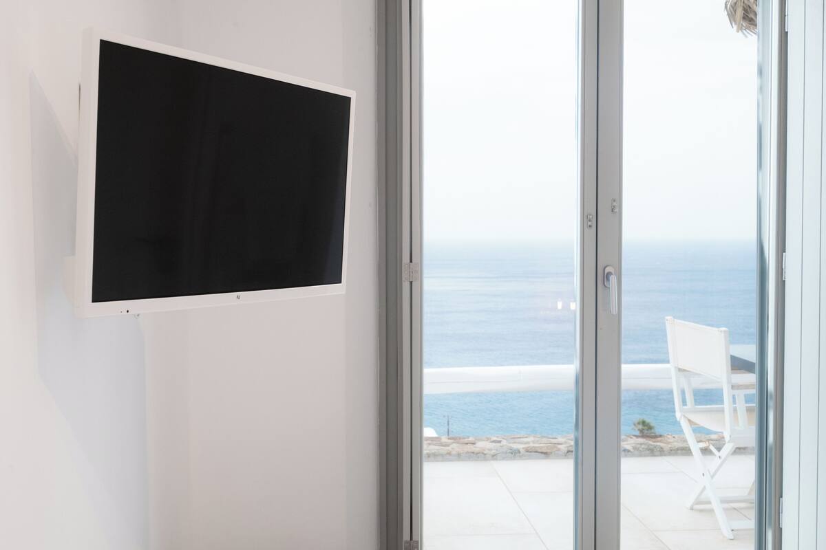 A room with a large TV mounted on a white wall, a sliding glass door leading to a balcony with a view of the sea.