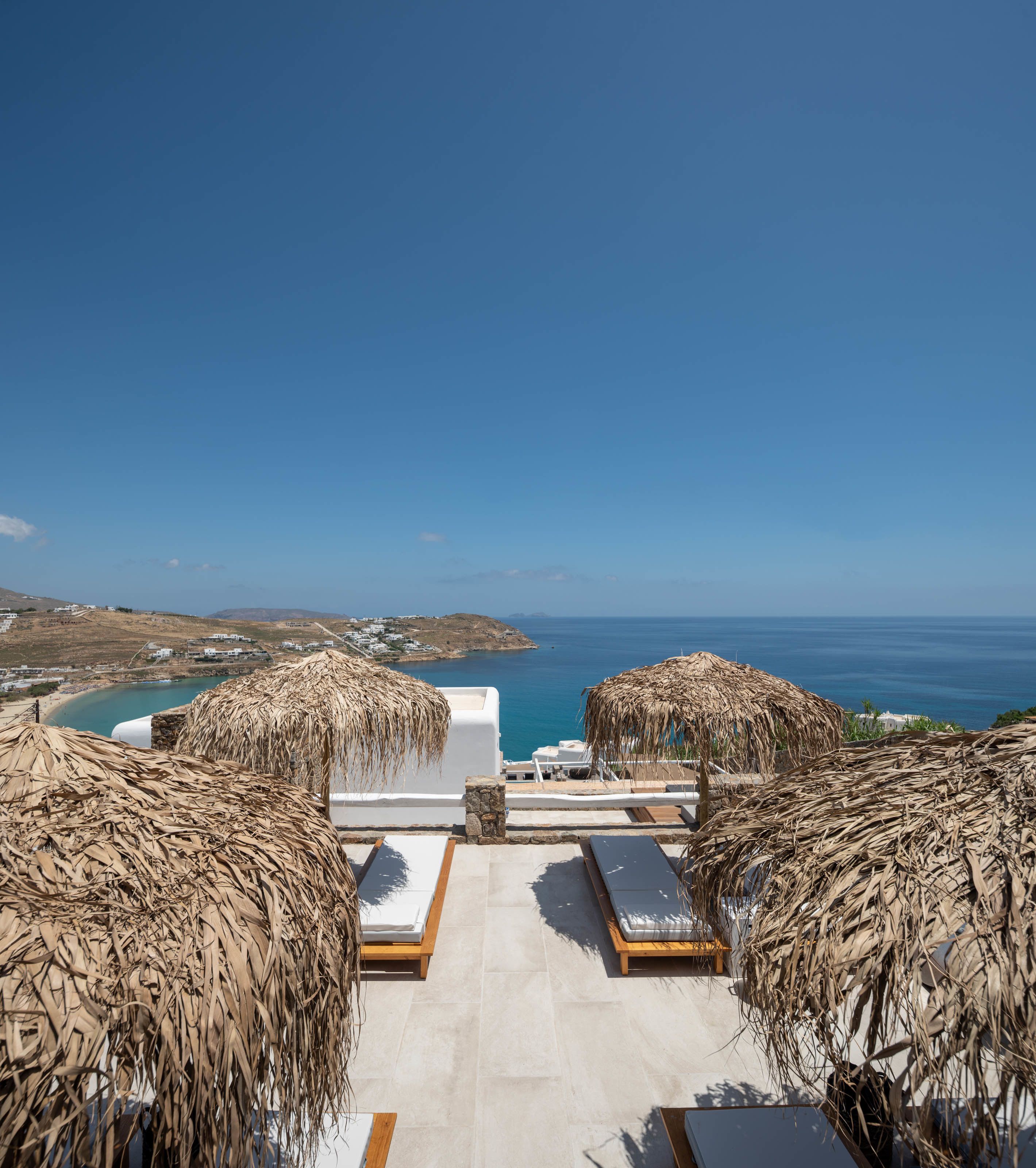 Una terrazza sul tetto con due lettini e un ombrellone di paglia, con vista sul paesaggio costiero e sul mare.