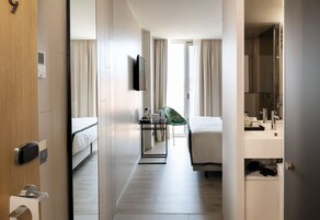 A hotel room with a bed, a desk, a chair, a mirror, and a bathroom with a sink and mirror.