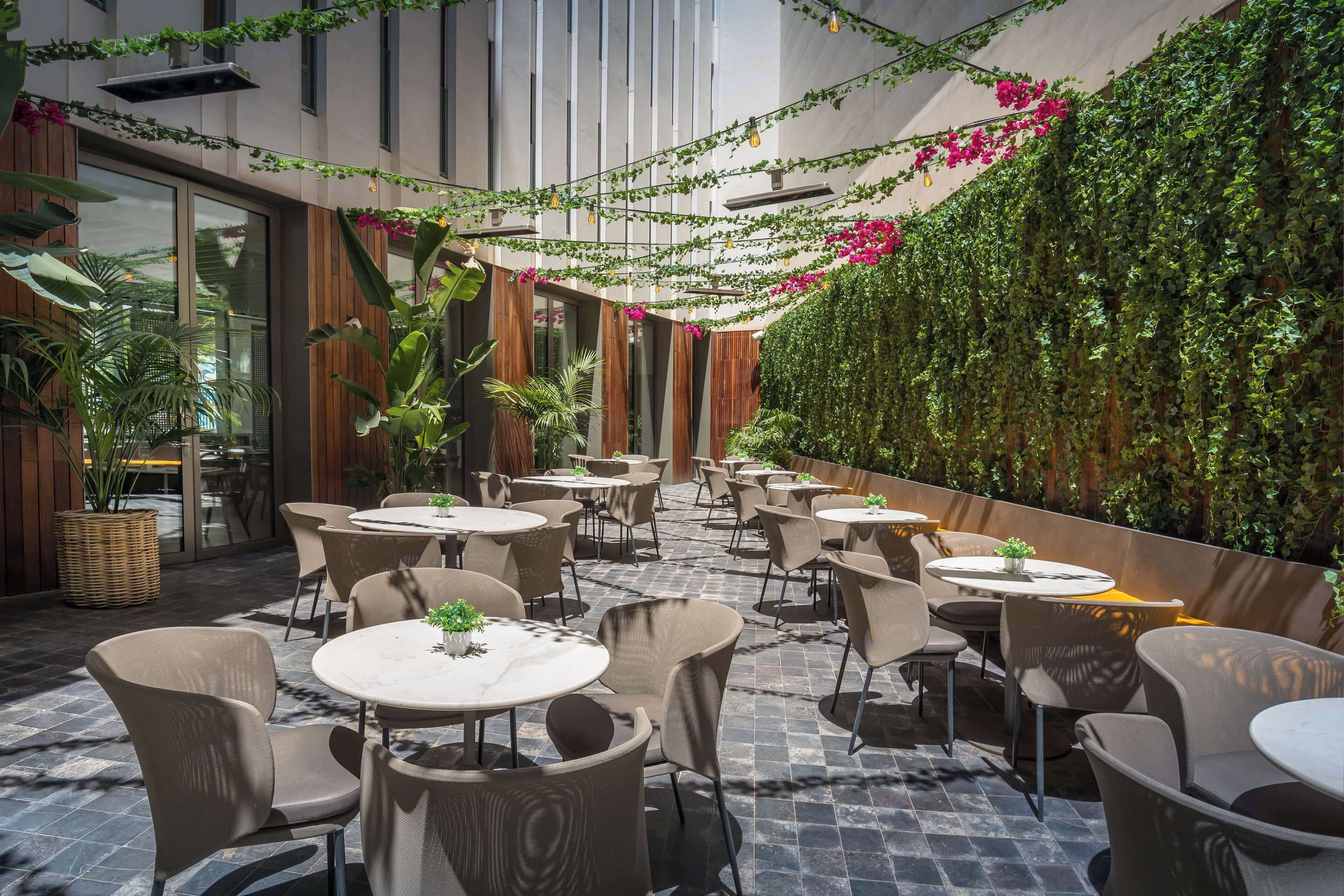 Un área de café al aire libre con mesas y sillas redondas, rodeada de abundante follaje y plantas.