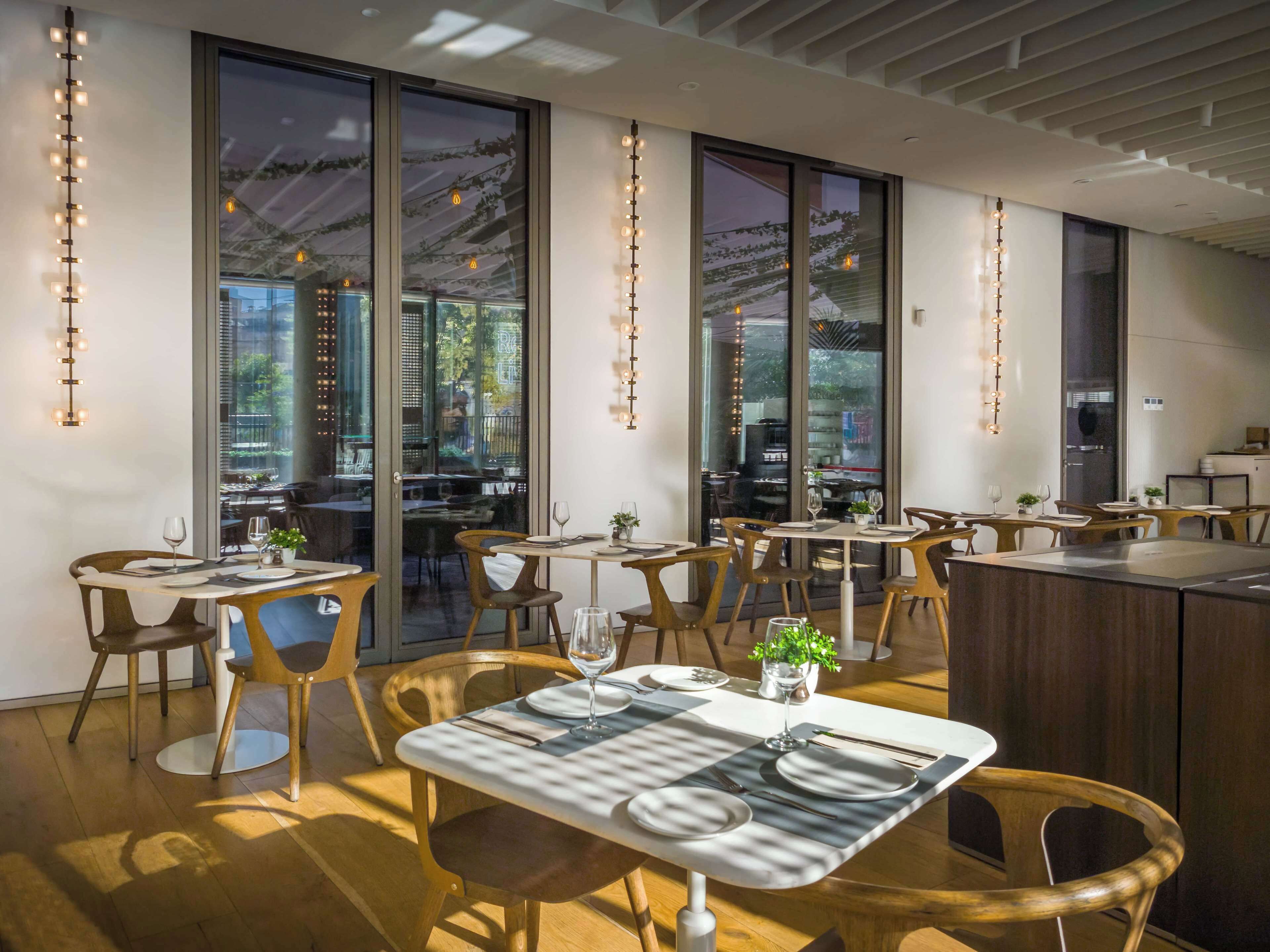 Moderni ravintola, jossa on suuret ikkunat, puiset tuolit ja ruokailuun katetut pöydät.