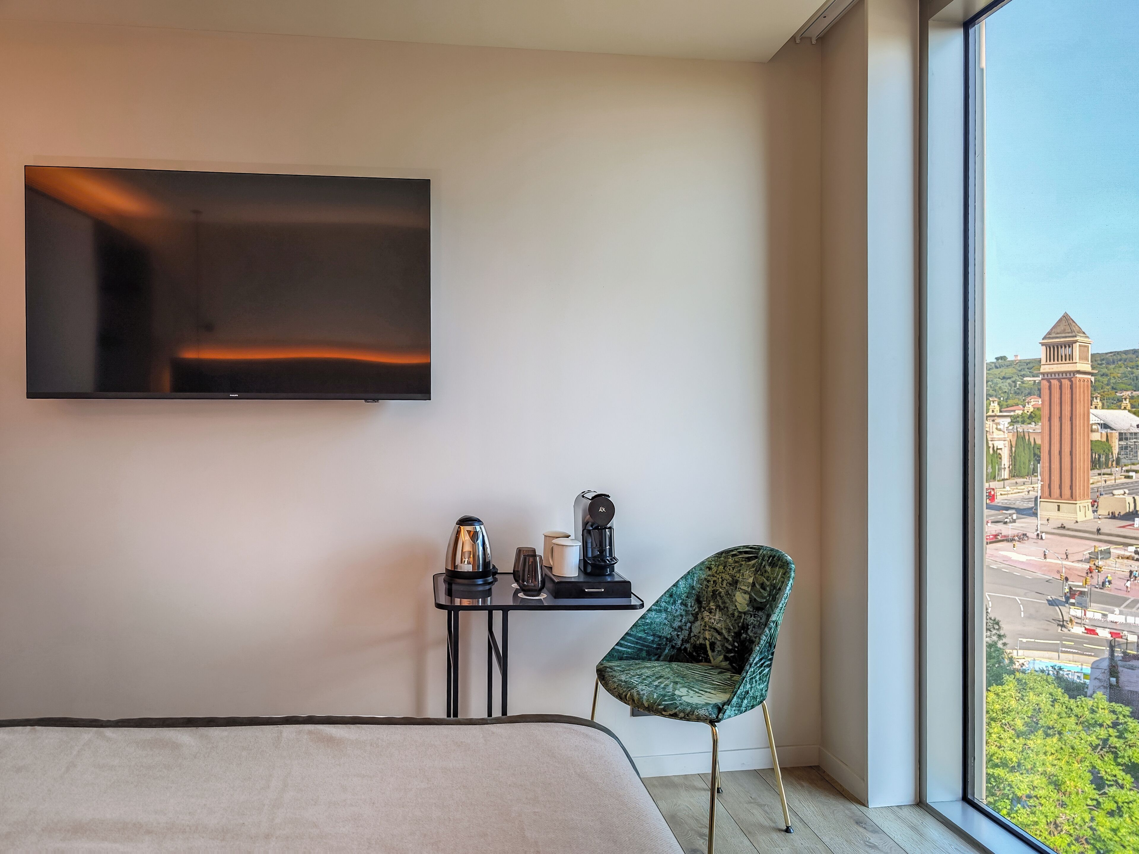A hotel room with a bed, a TV mounted on the wall, a small table with a kettle and cups, and a chair.