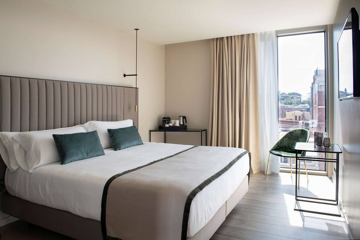 Quarto de hotel moderno com uma cama grande, uma mesa pequena com uma cadeira e vista para a cidade pela janela.