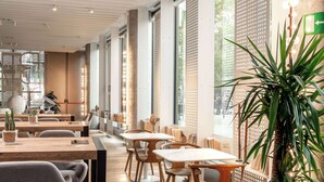 Un interior de cafetería moderna con mesas y sillas de madera, ventanas amplias y una planta en maceta.