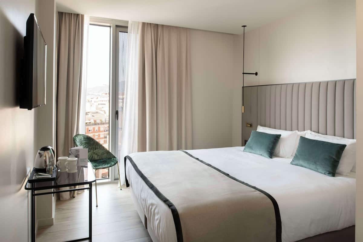 Quarto de hotel com cama, televisão, secretária com chaleira e chávenas, e uma cadeira.