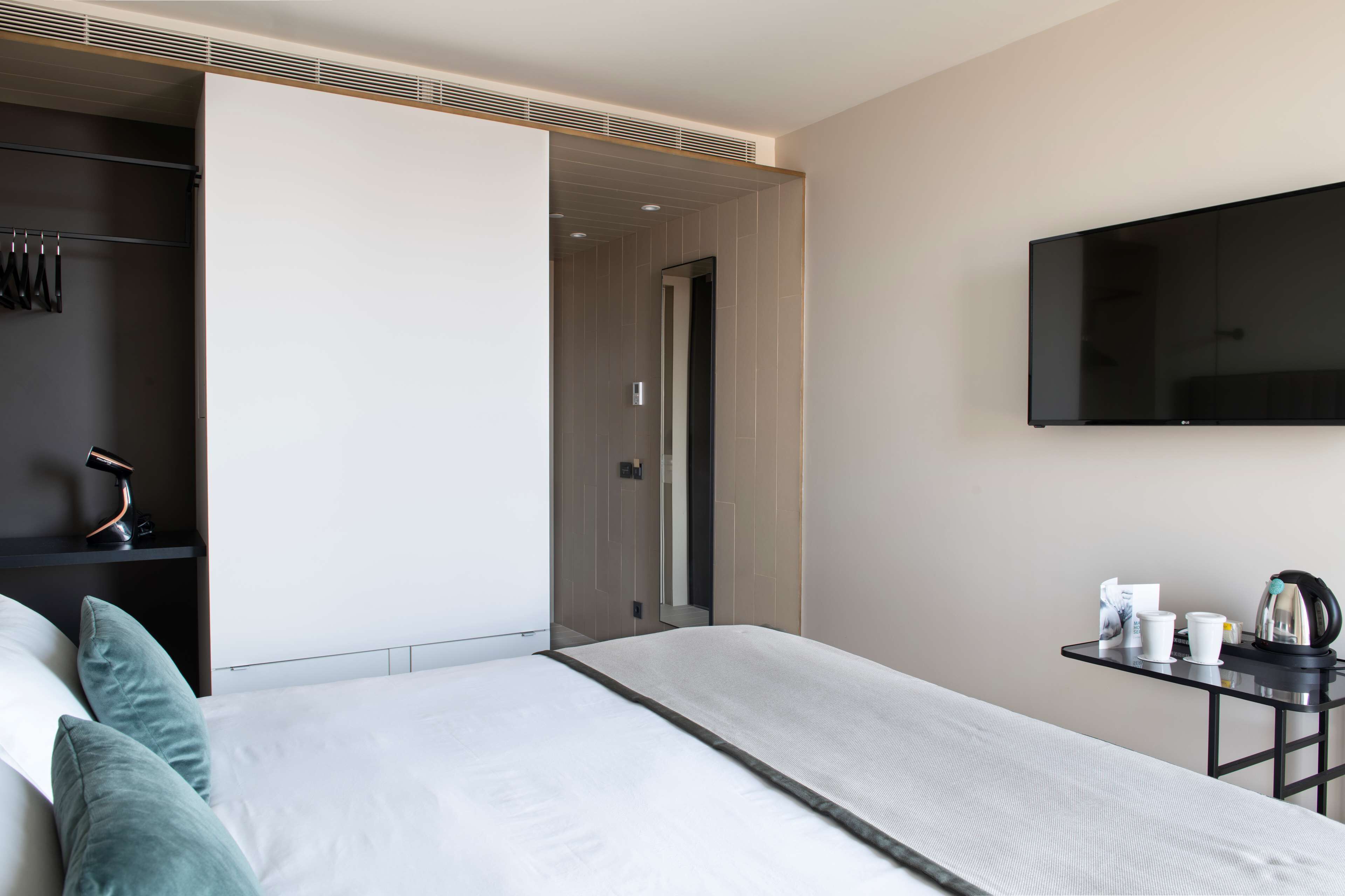 A modern hotel room with a bed, a TV mounted on the wall, and a small table with a kettle and cups.