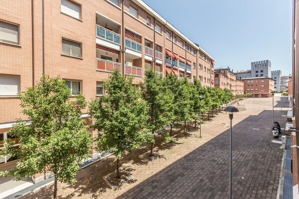 A multi-story brick building with balconies, a paved pedestrian area, and trees lining the street.