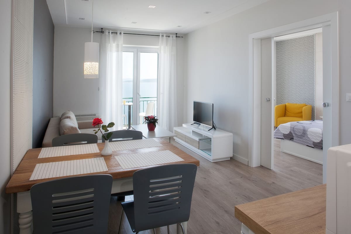 A modern living room with a dining area, a TV on a white stand, and a balcony with a yellow chair.