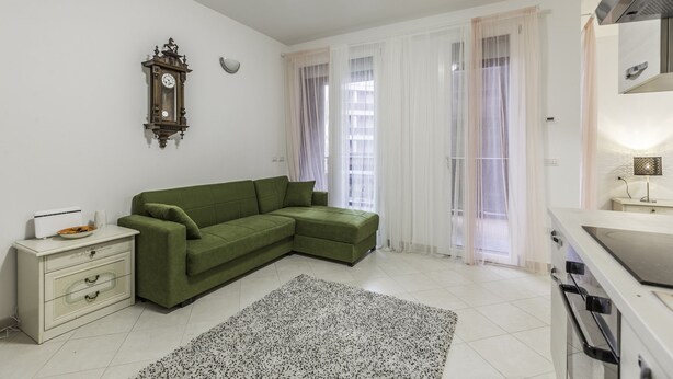 Ein modernes Wohnzimmer mit einem grünen Ecksofa, einer weißen Kommode, einer Wanduhr und einer Küchenzeile mit Herd.