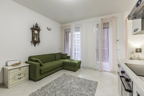 A modern living room with a green sectional sofa, a white dresser, a clock on the wall, and a kitchen area with a stove. - Appartamento Freud 8 (Bologna)