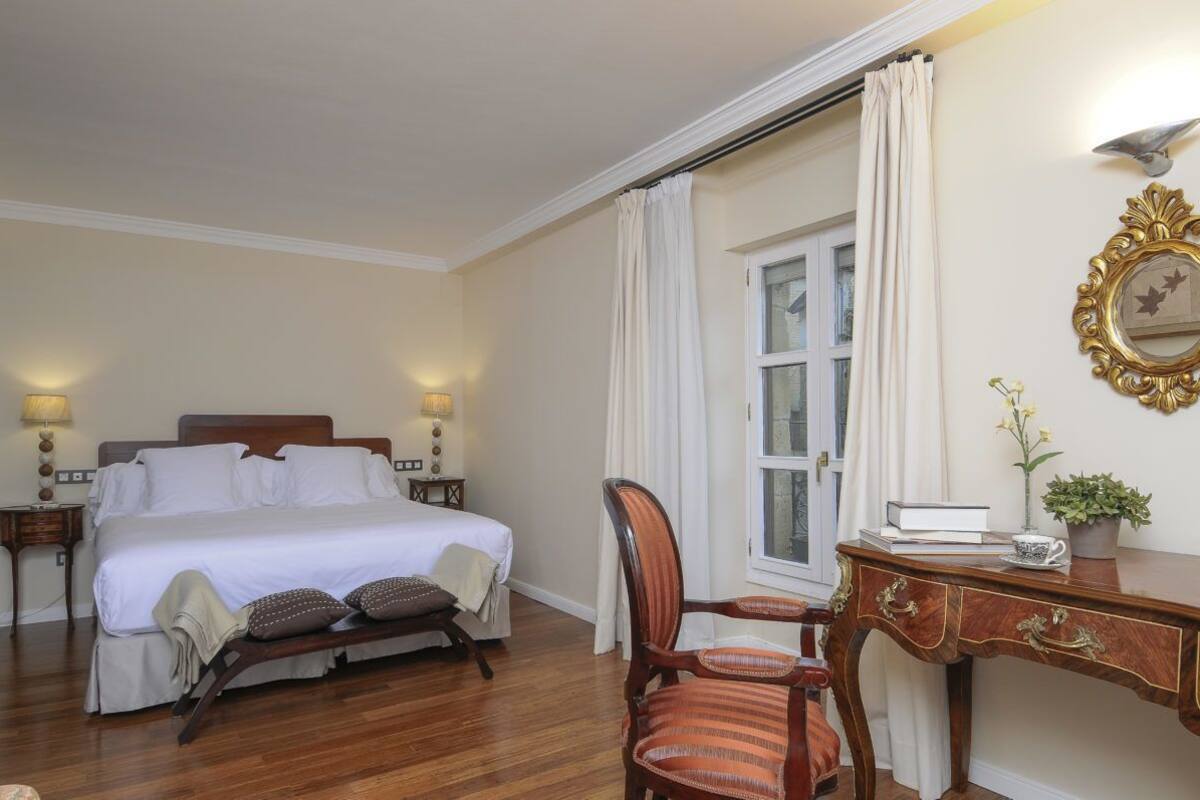 Une chambre d’hôtel avec un grand lit, un bureau avec une chaise, une fenêtre avec des rideaux, un miroir fixé au mur et une petite table avec une plante.