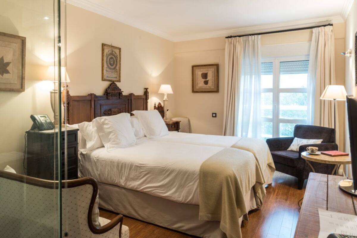 Une chambre d’hôtel avec un lit, un bureau, une chaise, une lampe et une fenêtre avec des rideaux.