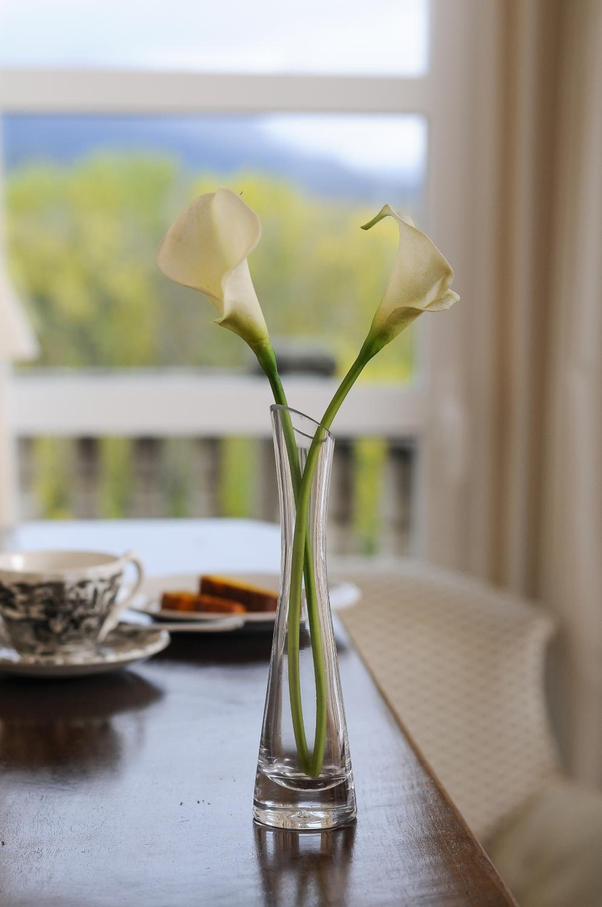 A clear glass vase with two white calla lilies on a wooden table by a window.