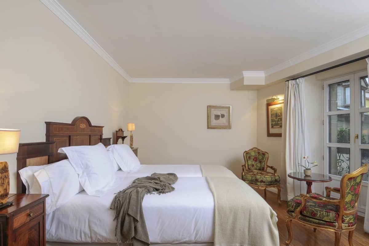 Une chambre d’hôtel avec un lit double, deux tables de chevet, une petite table d’appoint, deux chaises, une petite table et une fenêtre avec des rideaux.