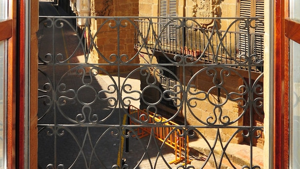 A view through a wrought iron gate into a narrow street with buildings featuring traditional architecture and balconies.