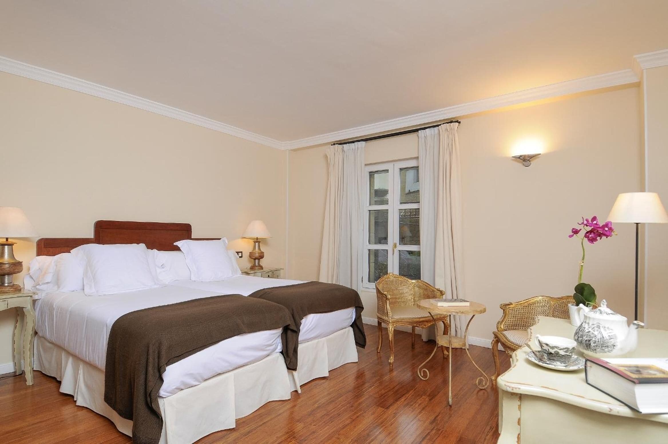 A hotel room with two beds, a sitting area with a table and chairs, a window with curtains, and a vase with flowers.