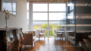 A spacious room with a large window offering a view of greenery and a distant landscape. There is a wooden dresser with ornate handles and a vase with flowers on top. The room has a modern design with a glass partition and a dark-colored sofa.