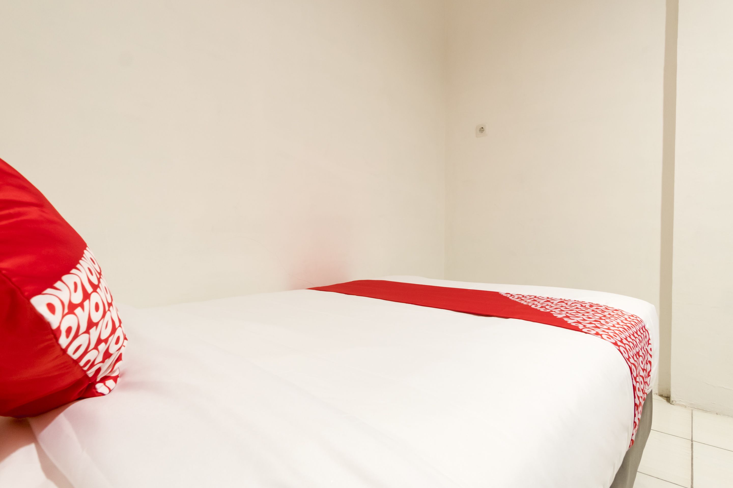 A bed with a white and red bedspread, a red pillow with white text, and a plain white wall.