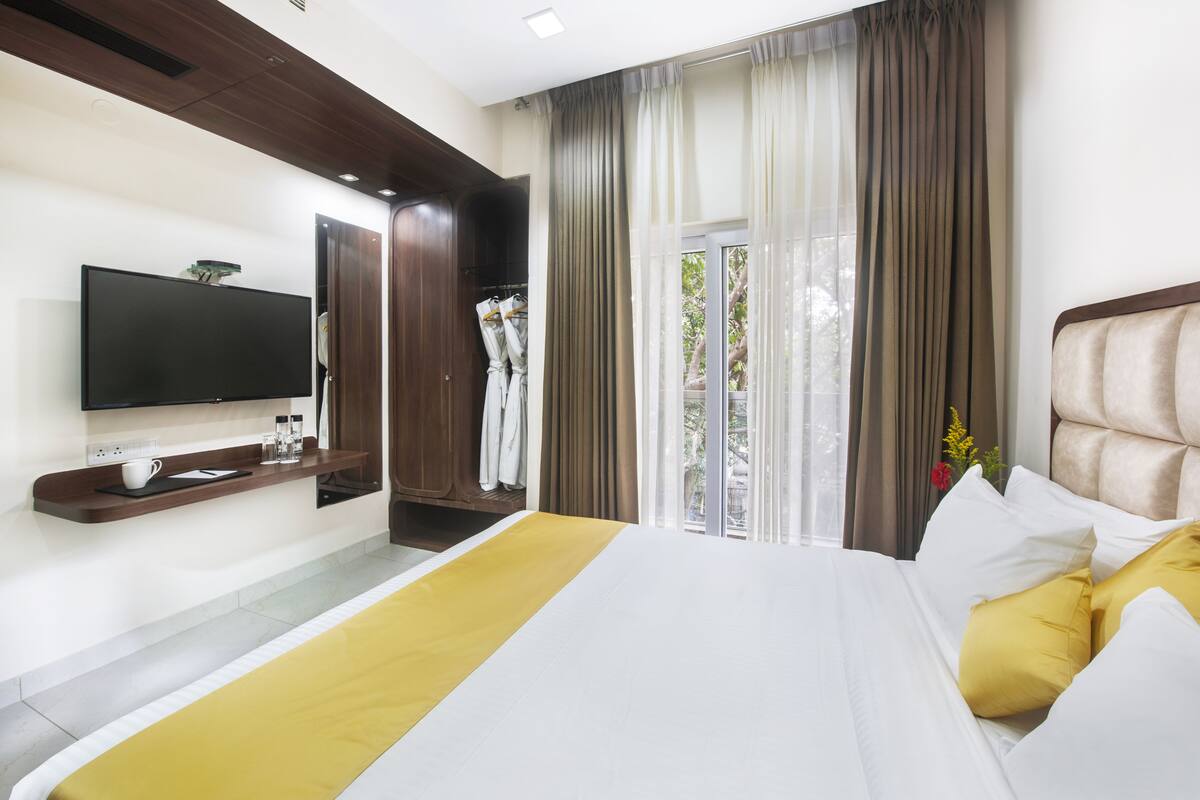 A modern hotel room with a large bed, a flat-screen TV, a bedside table with a cup and glasses, and a window with curtains.