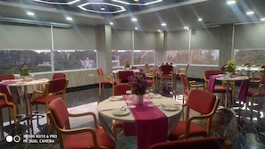 Een restaurant met ronde tafels en rode stoelen, grote ramen met uitzicht op de stad, en een modern plafond.