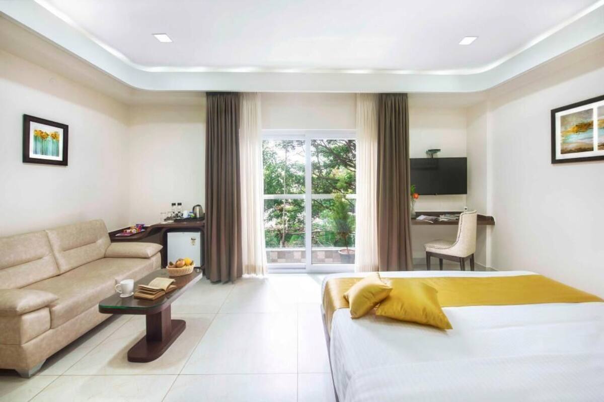 A hotel room with a sofa, a bed with yellow pillows, a desk with a TV, and a large window with curtains.
