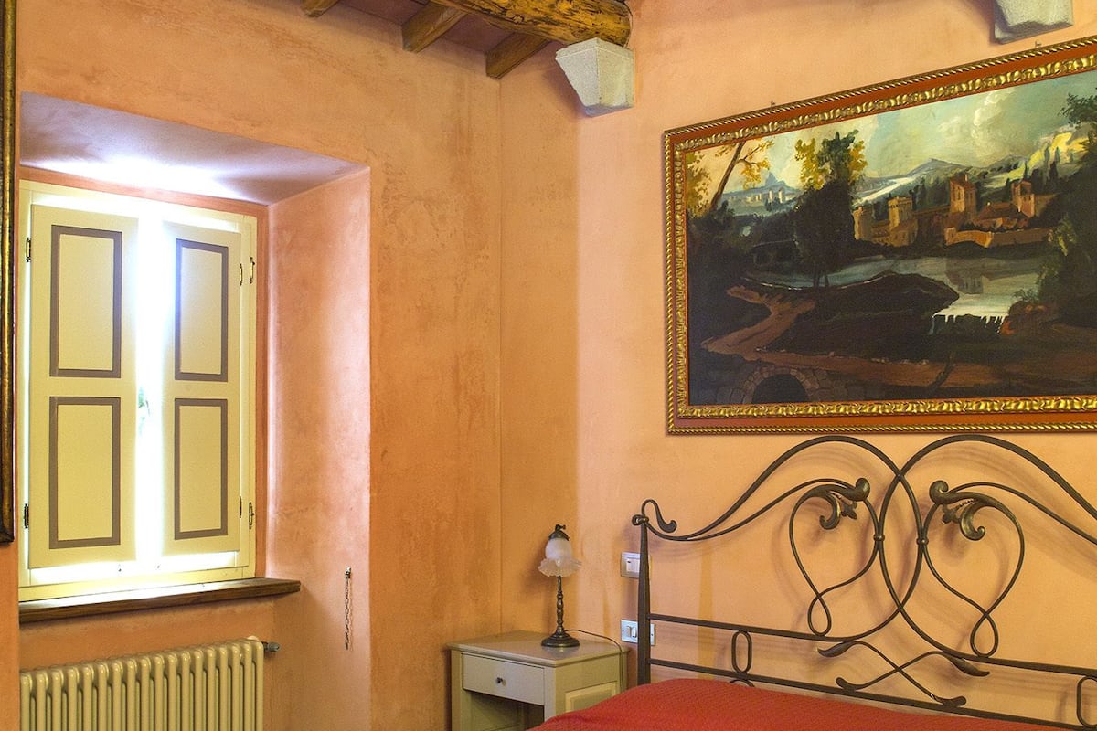 Ein Schlafzimmer mit Holzbalkendecke, einem Bett mit roter Bettdecke, einem Nachttisch mit Lampe und einem Gemälde an der Wand.