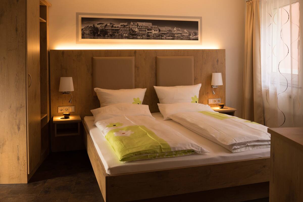Una habitación de hotel con cabecera de madera, una cama con ropa de cama blanca y detalles verdes, dos lámparas de noche, un cuadro enmarcado de una vista urbana sobre la cama, una botella de vino y una copa sobre una mesita de madera, y una ventana con cortinas.