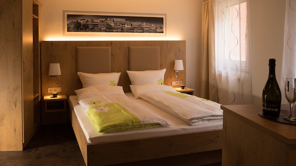 A hotel room with a wooden headboard, a bed with white bedding and green accents, two bedside lamps, a framed cityscape picture above the bed, a bottle of wine and a glass on a wooden side table, and a window with curtains.