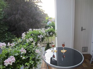 A round table with two wine glasses on a balcony with a view of a garden and a white door.