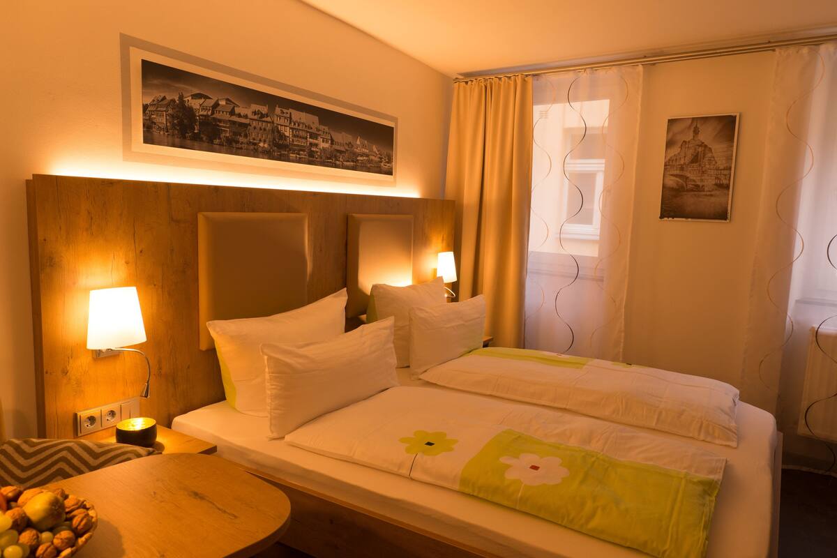Habitación de hotel con cabecera de madera, dos camas con ropa blanca, una mesita de noche con lámpara, una pequeña mesa con fruta y un cuadro en la pared.