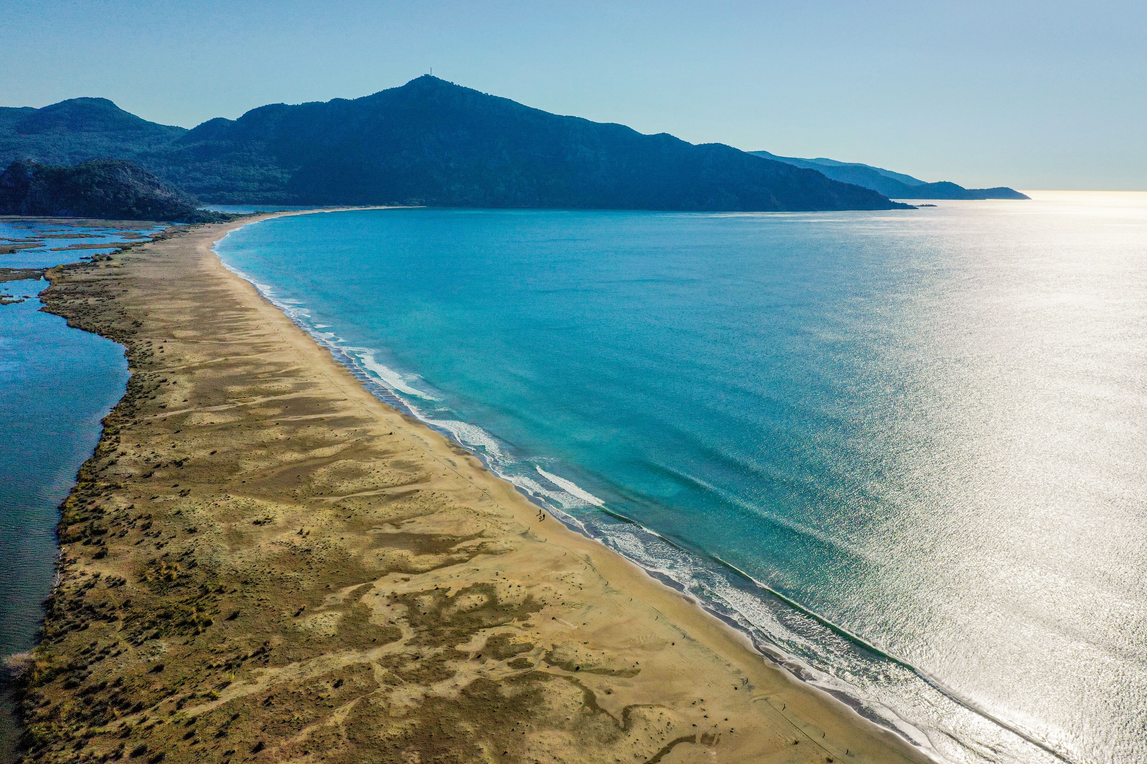 An aerial view of a beach with clear blue waters, a sandy shore, and mountains in the background.