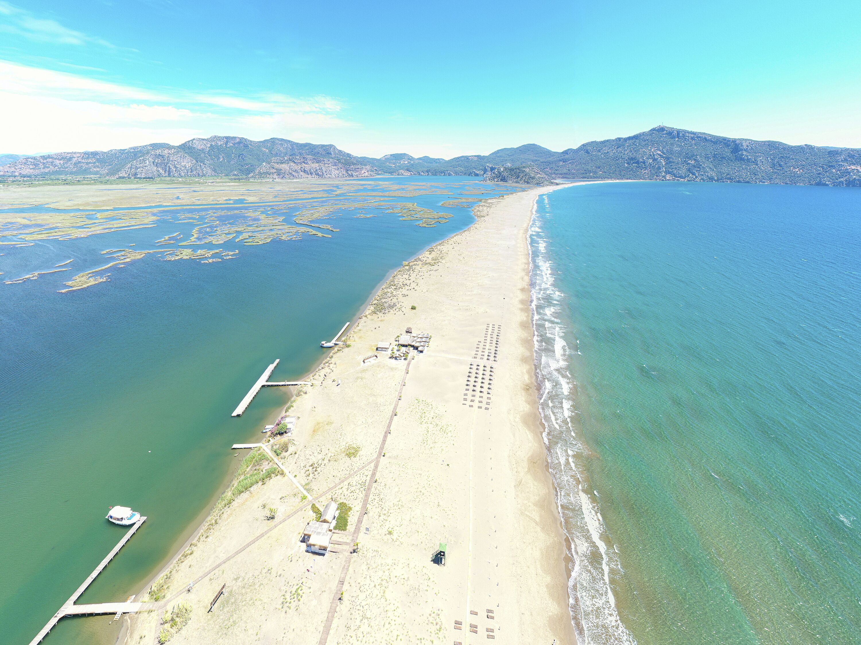 Vista aérea de una playa con un muelle largo que se adentra en el agua, cielo despejado y montañas a lo lejos.