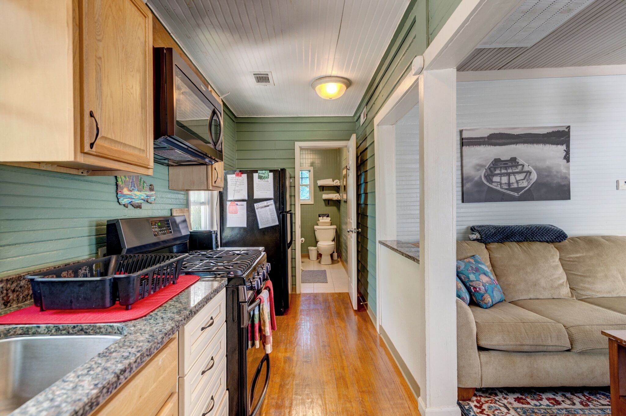 A compact kitchen with a stove, refrigerator, and wooden cabinets. A hallway leads to a living area with a couch and a rug.