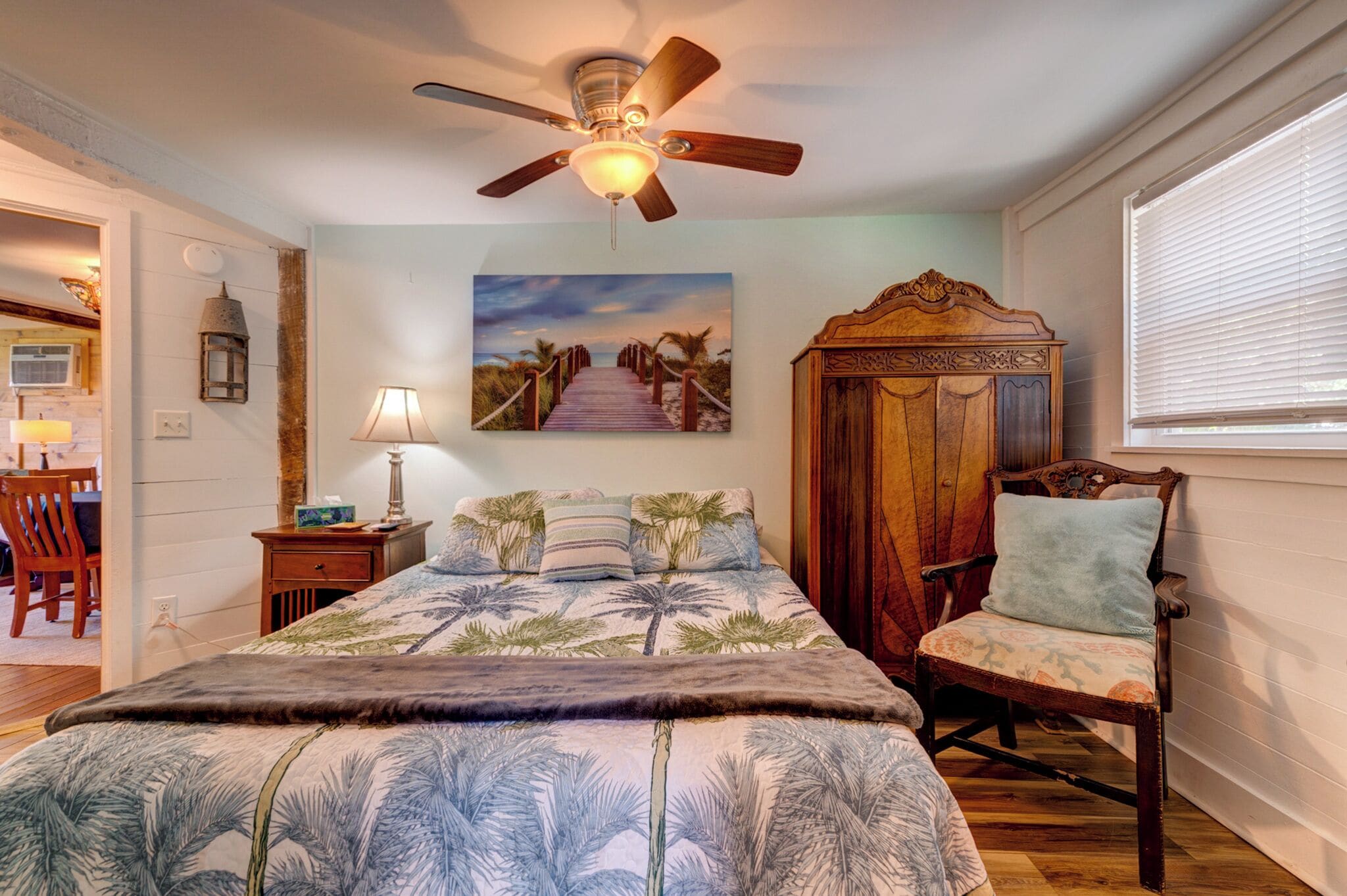 Une chambre à coucher avec un lit, une chaise en bois, un ventilateur de plafond, une table de chevet, une lampe, un tableau au mur et une fenêtre avec des stores.